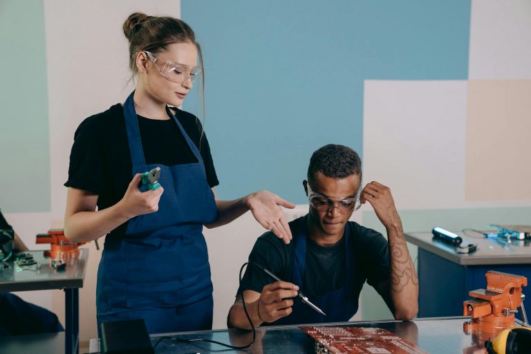 Two technicians working together on electronics with safety gear in a workshop.